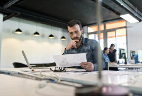 Man looking at document next to open laptop at desk in modern office