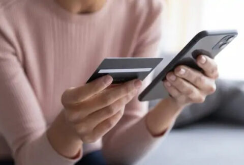 Woman sitting on sofa holding up bank card next to smartphone