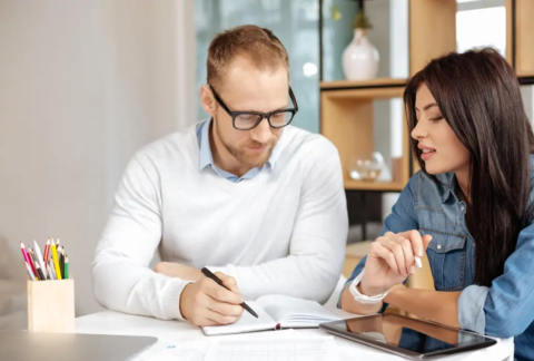 Two businesspeople sitting at table with tablet looking at open notebook