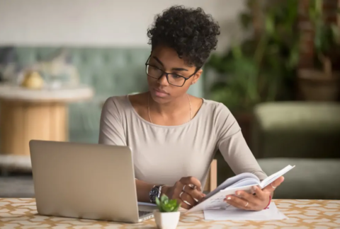 Businesswoman at table with open notebook in hands and looking at laptop to check National Insurance record