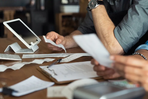Two business owners at table holding receipts and using tablet to calculate payments