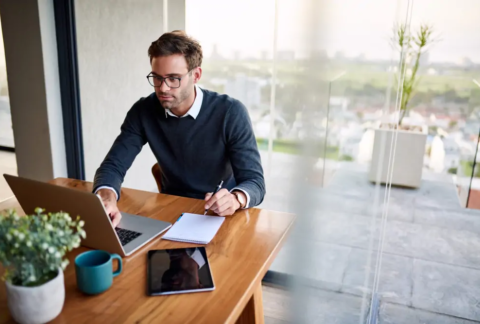 Businessman at desk holding pen over notebook and using laptop to calculate VAT
