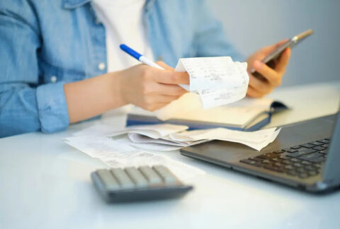 Businessperson holding smartphone in one hand and receipts and a pen in the other over an open account book and laptop