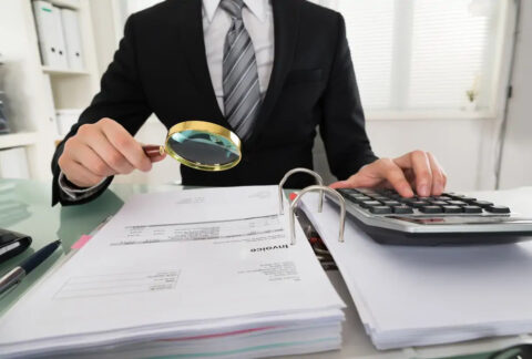 Businessman at desk using calculator with one hand and holding magnifying glass over invoice document in ring binder in the other to investigate business fraud