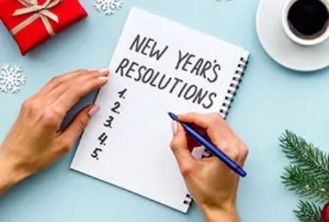 Hands writing list of New Year's resolutions in notebook surrounded by paper snowflakes, a miniature bauble, a miniature present, a fir branch and a cup of coffee