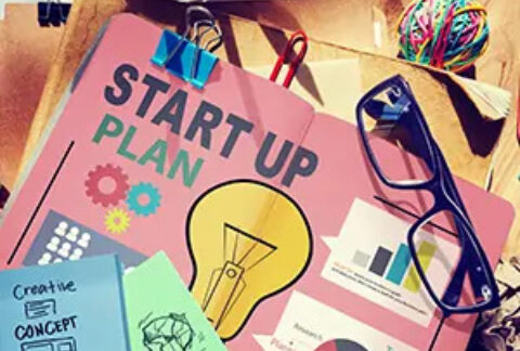 Colourful document titled 'start up plan' with pictures of a lightbulb, cogs and graphs on cover, on desk surrounded by post-it notes, elastic bands, paperclips, and a pair of glasses