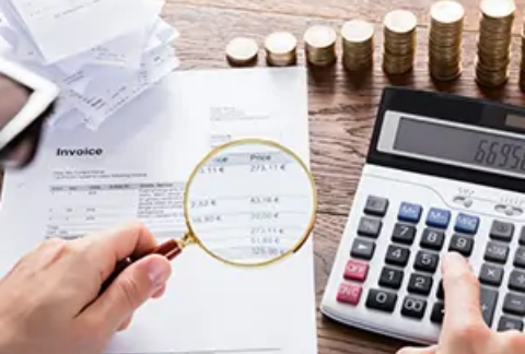 Person carrying out tax investigation at desk with one hand using calculator and the other holding magnifying glass over invoice