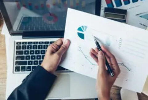 Businessperson holding pen over graph document above open laptop