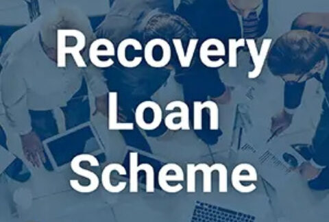 Businessmen standing around table discussing papers in meeting overlayed with text reading 'recovery loan scheme'