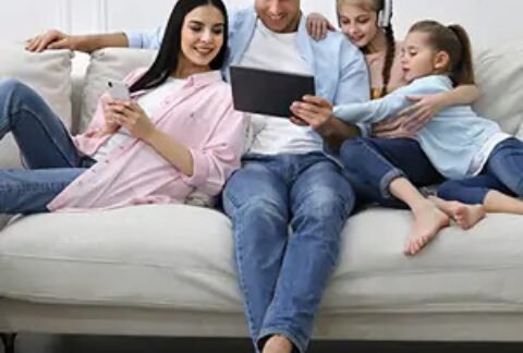 Young family sitting on sofa in home all looking at tablet that father is holding and smiling