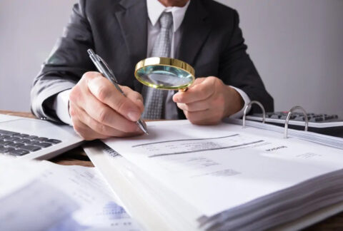 Businessman at desk holding pen and magnifying glass over invoice in ringbinder