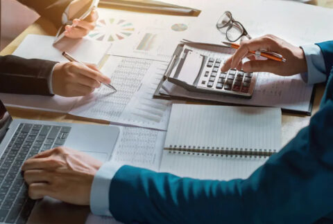 Two businesspeople sitting opposite each other at desk, one using a laptop and calculator and the other holding a smartphone and writing on a document