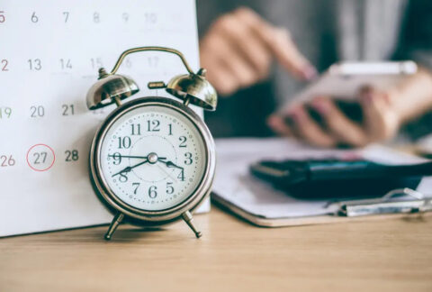Alarm clock in front of calendar next to businesswoman using smartphone in front of clipboard and calculator