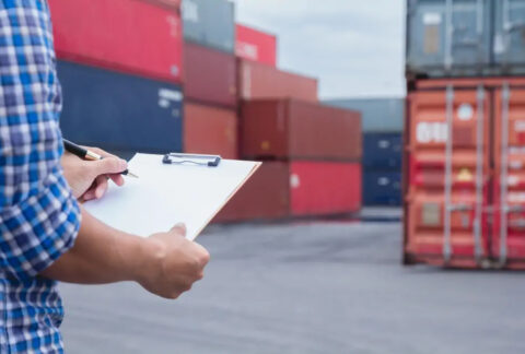 Man writing on clipboard standing in front of shipping containers