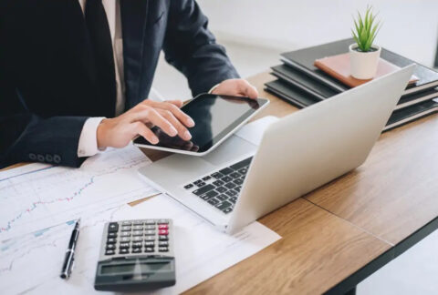 Businessman using tablet in front of open laptop next to calculator and graph documents
