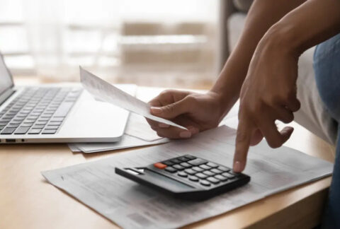 Woman holding paper in one hand and using calculator with the other next to open laptop