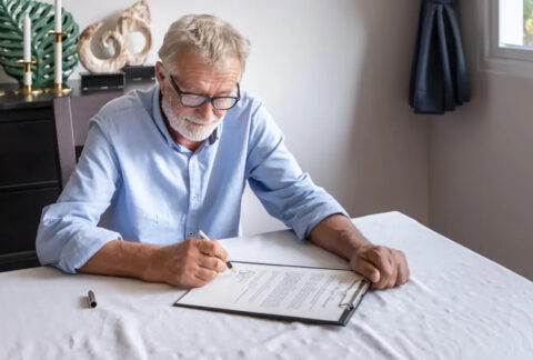 Elderly man at table signing form on clipboard
