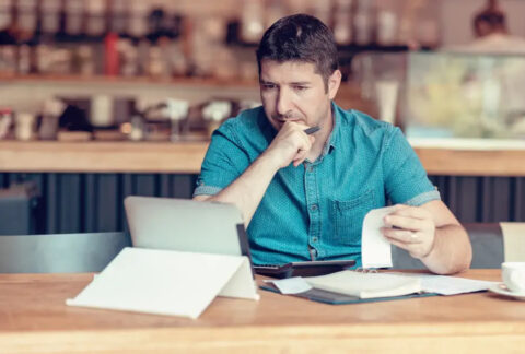 SME owner making calculations with receipts, tablet and calculator at table in cafe