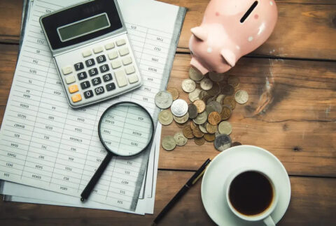 Calculator and magnifying glass on top of document, next to piggybank, pile of coins, and cup of coffee on wooden table