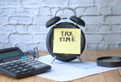 Alarm clock on desk on top of HMRC document and next to calculator and magnifying glass with post-it note reading 'tax time' over face
