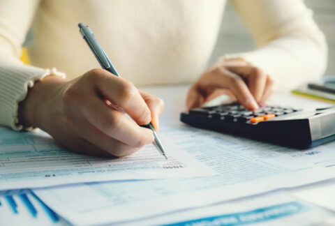 Businesswoman using calculator in one hand and filling in tax form with pen with the other