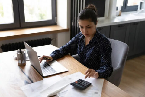 Businesswoman using calculator with one hand and laptop with the other to calculate dividends and salary