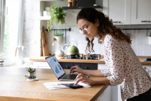 Businesswoman standing in kitchen using calculator and laptop on island to calculate year-end taxes