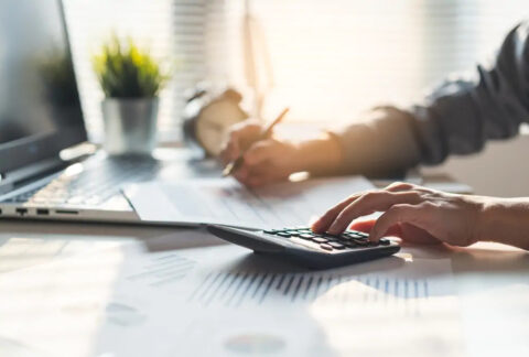 Businessman making tax calculations using calculator with one hand and writing on document on top of laptop with the other