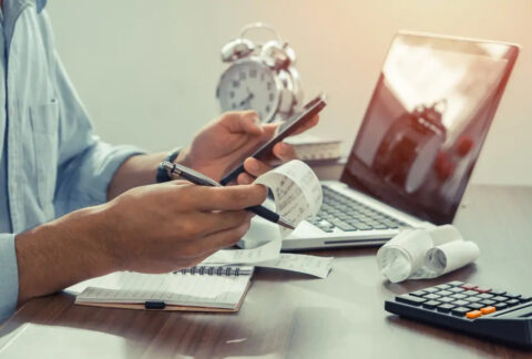 Businessman at desk holding smartphone in one hand and pen and receipts in the other