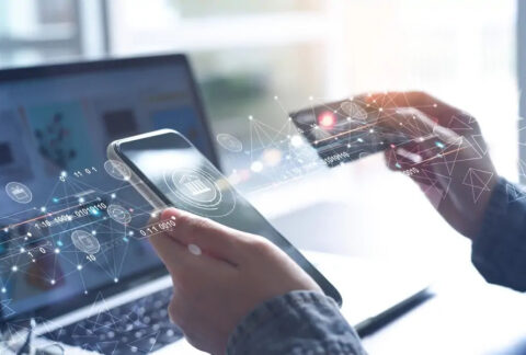 Businessperson using online banking app on smartphone and holding bank card above open laptop, overlaid with computer code images