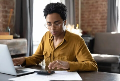 Businesswoman at desk using calculator with one hand and laptop with other