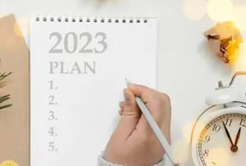 Woman's hand holding pencil over open notebook with '2023 business plan' and a list of numbers 1-5 written in it