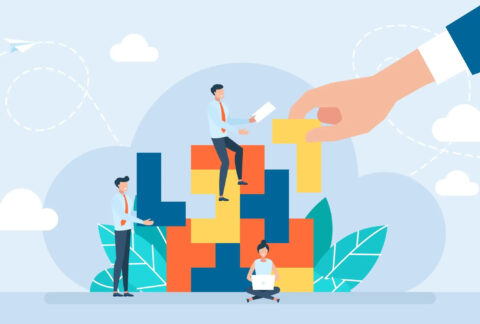 Cartoon of hand placing piece into Tetris-style puzzle with miniature businesspeople sitting and standing around puzzle with a laptop and document