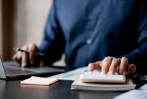 Businessman at desk using laptop with one hand and calculator with the other