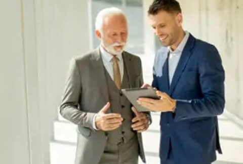 Younger businessman standing talking to elderly businessman and holding tablet that they are both looking at