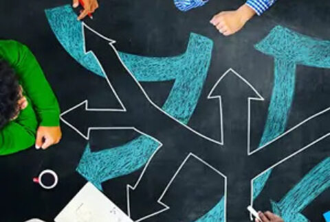 Businesspeople in meeting sitting around chalk board-style desk with arrows pointing in different directions drawn on it