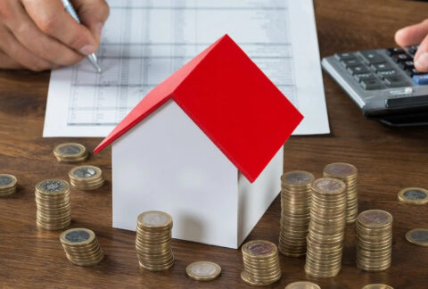 Businessman doing calculations beside a model house and stacks of coins