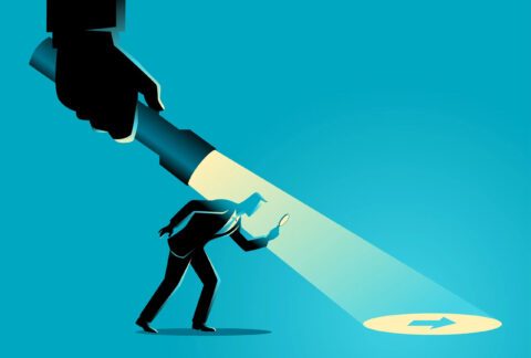 Cartoon of a businessman looking through a magnifying glass at an arrow on the floor that is illuminated by a giant torch held from above by a giant hand