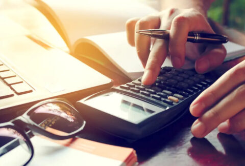 Businessperson at desk holding pen and using calculator to work out debt sums