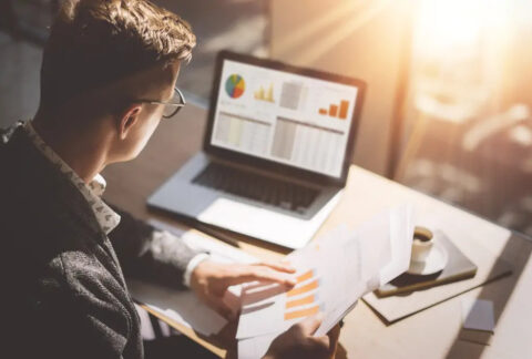 Businessman at desk looking at laptop screen and holding document displaying annual accounts