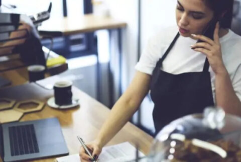 Barista behind coffee bar on the phone noting things down on a document next to an open laptop