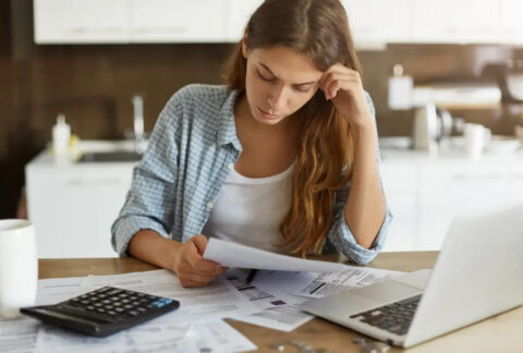 Woman reading document in front of laptop, calculator and more documents on desk